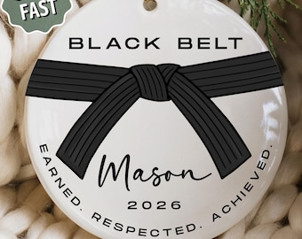 May include: A round white ceramic ornament featuring a black martial arts belt graphic across the center. The text reads BLACK BELT, Mason, 2026, and EARNED. RESPECTED. ACHIEVED. A gold string is attached to the top for hanging purposes.