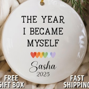 May include: White ceramic ornament with gold string, featuring the text "THE YEAR I BECAME MYSELF" in black. Below, it reads "Sasha 2025" with rainbow hearts. The ornament is round and has a glossy finish.