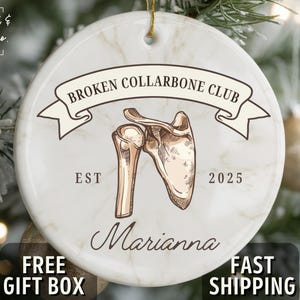 May include: A round ceramic ornament with a marble effect background. The ornament features a graphic of a broken collarbone and the text "BROKEN COLLARBONE CLUB" above it. The name "Marianna" is written below the graphic. The ornament is for the year 2025.