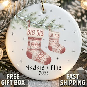 May include: White ceramic Christmas ornament with a gold string for hanging. The ornament features two stockings, one labeled "BIG SIS" and the other "LIL SIS". The names "Maddie + Ellie" and the year "2025" are printed below the stockings.