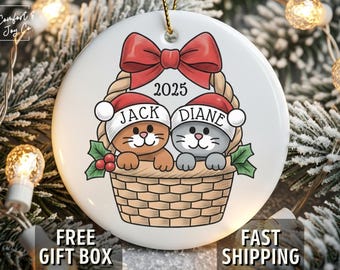 May include: A white ceramic Christmas ornament featuring two cartoon cats in a basket, wearing Santa hats. The ornament has a red bow, the year 2025, and the names "Jack" and "Diane". The ornament is hanging on a gold string.