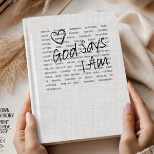 Peut inclure: Journal ivoire avec l'inscription "God Says I Am" dans une police manuscrite. La couverture pr&eacute;sente un motif imprim&eacute; mat et une texture en faux lin. Les mots "Chosen, Saved, Strong, Loved" sont imprim&eacute;s autour du texte principal.