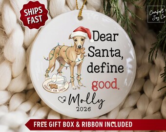 May include: A round white ceramic ornament featuring a tan dog wearing a Santa hat and tangled in colorful Christmas lights. The dog holds a gingerbread cookie in its mouth. Text reads: Dear Santa, define good. Molly 2026.