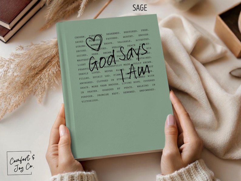 Peut inclure: Journal vert sauge avec l'inscription "God Says I Am" et d'autres affirmations positives. Le journal comporte un gribouillage de c&oelig;ur et est ouvert, montrant le texte. Le journal fait partie de la collection Comfort & Joy Co.