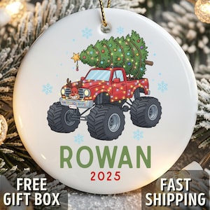 May include: White ceramic Christmas ornament featuring a red monster truck carrying a decorated Christmas tree. The ornament has the name "ROWAN" and the year "2025" printed on it. The truck has reindeer antlers and is decorated with lights.