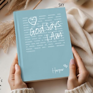 Peut inclure: Un journal bleu clair avec l'inscription blanche "God Says I Am". Il comporte un c&oelig;ur et le pr&eacute;nom "Harper". Le journal est tenu par une personne.