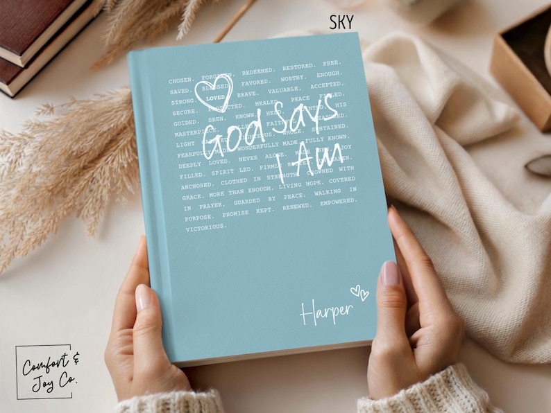 Peut inclure: Un journal bleu clair avec l'inscription blanche "God Says I Am". Il comporte un c&oelig;ur et le pr&eacute;nom "Harper". Le journal est tenu par une personne.
