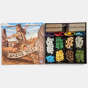 Puede incluir: Una caja de juego de mesa Skara Brae con la ilustración de una persona con una lanza, junto a una caja abierta que contiene pilas de cartas y varios marcadores de madera de colores en bandejas de plástico negro, incluyendo blanco, verde, beige y azul.