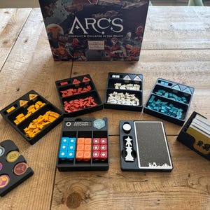 Op de afbeelding: Een set zwarte plastic organizers voor het bordspel "Arcs: Conflict & Collapse in the Reach". De organizers bevatten compartimenten voor verschillende gekleurde plastic onderdelen, dobbelstenen en kaarten. De speldoos is ook zichtbaar op de achtergrond.