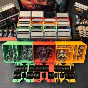 Könnte beinhalten: Ein Brettspiel-Organizer mit mehreren Fächern und Tabletts. Der Organizer ist schwarz, grün, orange und gelb. Er enthält Karten, Miniaturen und Spielsteine. Die Spielbox mit dem Titel "Nemesis: Retaliation" ist im Hintergrund sichtbar.