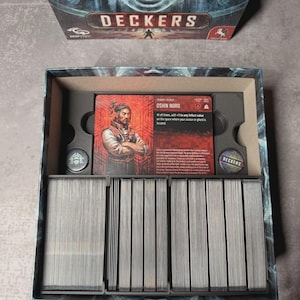 May include: An open board game box, "Deckers", displaying game cards and components. The central card features a portrait of a man named Oshin Nord. The box has a dark blue and grey colour scheme.