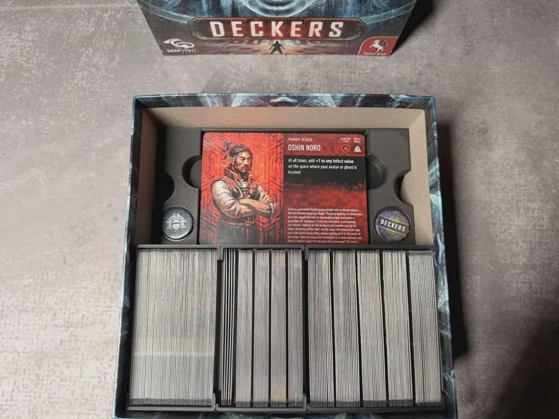 May include: An open board game box, "Deckers", displaying game cards and components. The central card features a portrait of a man named Oshin Nord. The box has a dark blue and grey colour scheme.