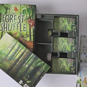 Pode incluir: Jogo de tabuleiro "Forest Shuffle" com tema florestal. A caixa apresenta um pica-pau, um esquilo, um veado e borboletas. No interior, há cartas com imagens de plantas e uma bandeja de armazenamento verde escuro.