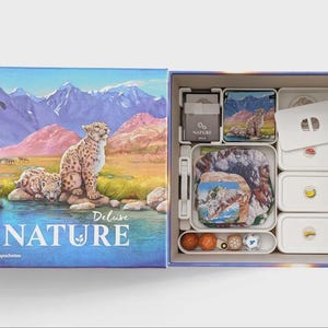 Könnte beinhalten: Geöffnete Deluxe Nature Brettspielbox mit Inhalt. Die Box zeigt Schneeleoparden an einem See mit Bergen. Im Inneren befinden sich Spielsteine und Aufbewahrungsbehälter. Die Box ist geöffnet und zeigt den Inhalt des Spiels.