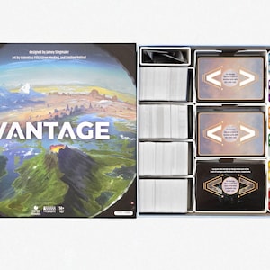 Op de afbeelding: Open Vantage bordspeldoos met spelcomponenten. De doosafbeelding toont een landschap met het woord "VANTAGE". Binnenin bevinden zich kaartenstapels, fiches en speelkaarten met tekst en symbolen.