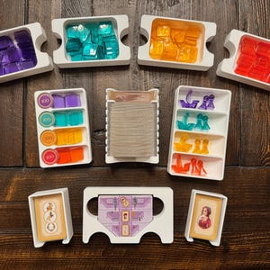 Puede incluir: Un conjunto de bandejas de plástico blanco con diversas piezas de juego de colores, incluyendo morado, azul, naranja y rojo. Las bandejas están etiquetadas con el número 100. También hay cartas y una pequeña figura.