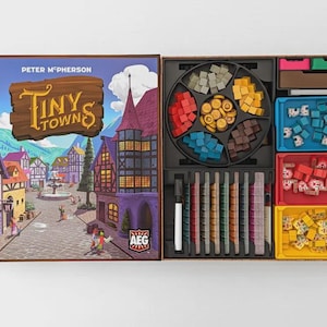 May include: Open board game box titled "Tiny Towns" by Peter McPherson. The box art depicts a colourful town scene. Inside, various game components are organised in trays, including wooden pieces in multiple colours and shapes, and game cards.