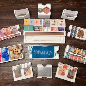 Puede incluir: Un organizador de juegos de mesa con varios componentes, incluyendo tarjeteros, bandejas para fichas y una bolsa azul con "SPEAKEASY" impreso. Las piezas del juego son de varios colores y formas, con denominaciones de 10 a 300.