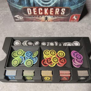 May include: A black game organiser with compartments filled with game pieces, including circular tokens in various colours and stacks of cards. The game box, labelled "DECKERS," is visible in the background.