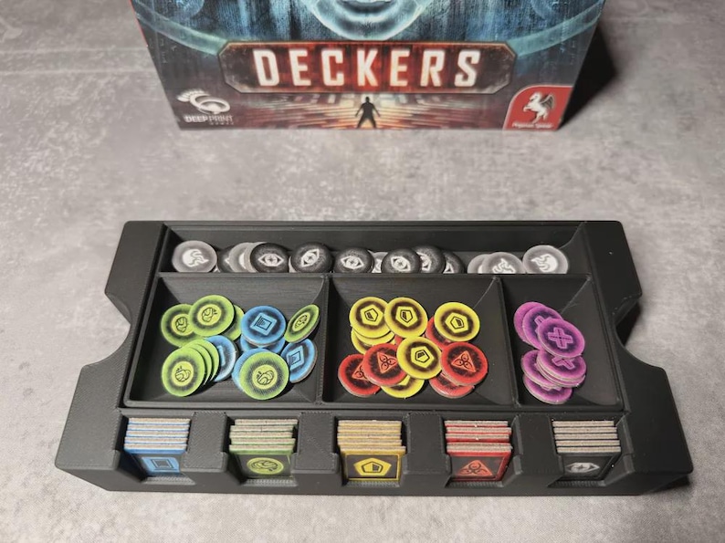 May include: A black game organiser with compartments filled with game pieces, including circular tokens in various colours and stacks of cards. The game box, labelled "DECKERS," is visible in the background.