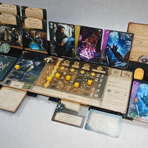 Puede incluir: Un organizador de juegos de mesa negro con varias cartas, fichas y un tablero de juego. Las cartas presentan arte y texto de fantasía. Se ven cubos amarillos y fichas circulares. El organizador está diseñado para guardar componentes del juego.