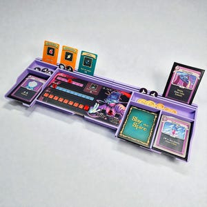 May include: A purple board game organizer with multiple compartments and card slots. The organizer holds game cards, tokens, and a game board with a colorful illustration. The game cards have various designs and text, including "Slay the Spire".