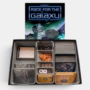 May include: A board game box for Race for the Galaxy featuring a space station illustration. The open box contains a grey plastic insert with compartments holding stacks of cards, orange hexagonal tokens, small white dice, and tan game cards.
