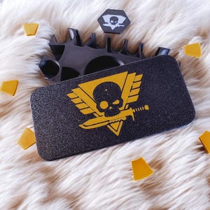 May include: A black box with a skull and crossbones logo on the lid. The logo is yellow and features a winged skull with a sword. The box is surrounded by small yellow triangular pieces.