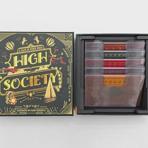 Op de afbeelding: Open bordspeldoos met de titel "HIGH SOCIETY" in gouden letters op een zwarte achtergrond. De doos is versierd met gouden illustraties van een zeilboot, vliegtuig en casinofiches. De spelcomponenten zijn netjes geordend.