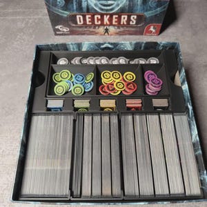 May include: An open board game box labelled "DECKERS". Inside are stacks of cards, and various coloured tokens in green, blue, yellow, and purple, plus silver tokens. The box is black with a blue and grey design.