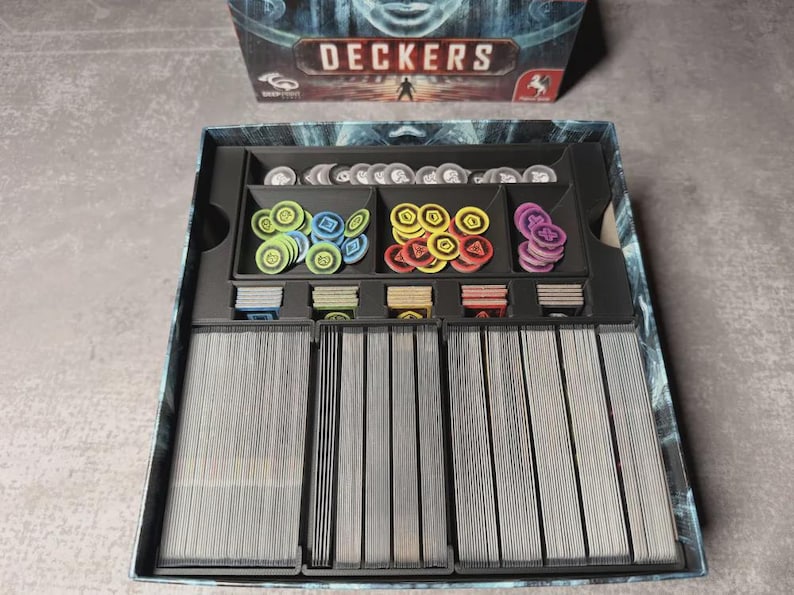 May include: An open board game box labelled "DECKERS". Inside are stacks of cards, and various coloured tokens in green, blue, yellow, and purple, plus silver tokens. The box is black with a blue and grey design.