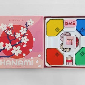 Puede incluir: Una caja de juego de mesa titulada Hanami Deluxe Edition de Reiner Knizia, con una cubierta rosa, un sol rojo y flores de cerezo blancas. La caja abierta contiene una bandeja de plástico blanca con fichas amarillas, azules, rojas y verdes, pequeñas piezas y cartas.