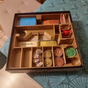 Può includere: Una scatola di legno marrone con un bordo nero e un interno dorato. La scatola ha diversi scomparti per riporre i pezzi del gioco, tra cui uno scomparto etichettato "Structure par Age". La scatola contiene anche una varietà di pezzi del gioco, tra cui monete, gettoni e carte.