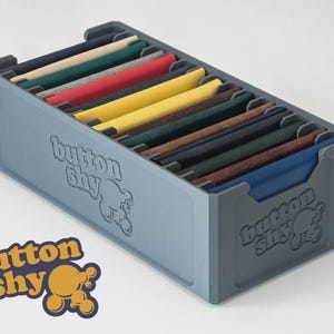 May include: A grey storage box with the text "button shy" embossed on the side, filled with fabric swatches in various colours. The swatches include white, green, red, yellow, and blue. The "button shy" logo is also visible.