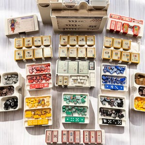 May include: A collection of colorful plastic storage containers with various compartments for organizing small game pieces. The containers are arranged in rows on a light wood surface.