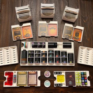 Op de afbeelding: Witte plastic opbergcontainers voor een bordspel. De containers zijn gevuld met verschillende spelstukken, waaronder kleine zwarte en witte tokens, rode en gele dobbelstenen en kleine gekleurde blokjes. De containers zijn gelabeld met de tekst "Lost Legacy".