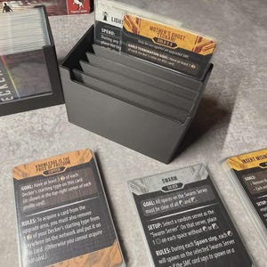 May include: A grey card holder containing game cards. The cards feature text and graphics, including "Mother's Ghost Tsunami" and "Knowledge is the Price of Freedom". The holder is on a light surface.