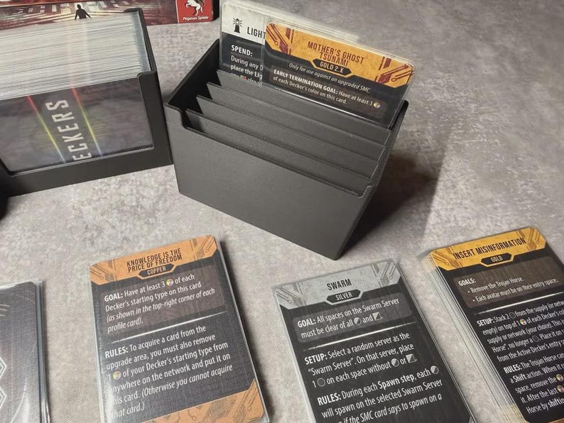 May include: A grey card holder containing game cards. The cards feature text and graphics, including "Mother's Ghost Tsunami" and "Knowledge is the Price of Freedom". The holder is on a light surface.