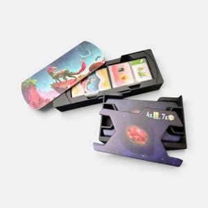 K&ouml;nnte beinhalten: Ein schwarzer Kartenorganizer aus Kunststoff f&uuml;r Brettspiele mit zwei F&auml;chern. Ein Fach enth&auml;lt einen Kartenstapel mit einer Weltraum-Illustration einer Kreatur, w&auml;hrend das andere ein violettes Galaxie-Design mit dem Aufdruck 4x und 7x zeigt.