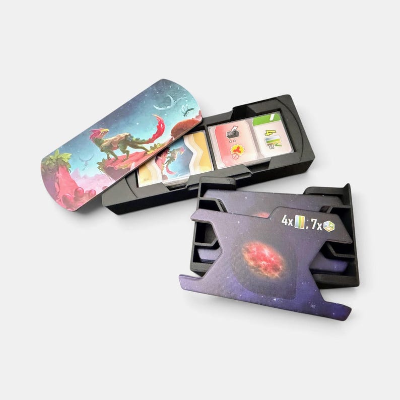 K&ouml;nnte beinhalten: Ein schwarzer Kartenorganizer aus Kunststoff f&uuml;r Brettspiele mit zwei F&auml;chern. Ein Fach enth&auml;lt einen Kartenstapel mit einer Weltraum-Illustration einer Kreatur, w&auml;hrend das andere ein violettes Galaxie-Design mit dem Aufdruck 4x und 7x zeigt.