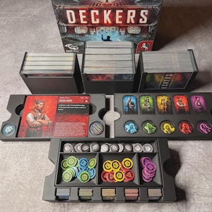 May include: A board game set with the title "DECKERS" on the box. The set includes card holders, card decks, character cards, and various game tokens in a black organizer. The tokens are in various colors, including blue, yellow, and purple.