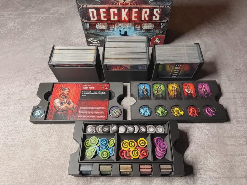 May include: A board game set with the title "DECKERS" on the box. The set includes card holders, card decks, character cards, and various game tokens in a black organizer. The tokens are in various colours, including blue, yellow, and purple.