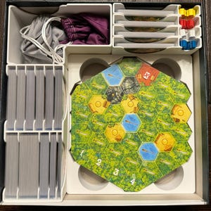 Puede incluir: Una caja organizadora blanca con compartimentos para guardar piezas de juego. La caja contiene un tablero de juego hexagonal con fichas verdes y marrones, un juego de cartas y una bolsa de fichas. La caja está abierta y el contenido es visible.