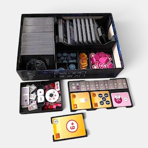 May include: A board game box containing organized card stacks, blue circular tokens, orange hexagonal tokens, and pink circular tokens with star icons. Two black trays hold white dice, small rectangular tiles, and square game cards.