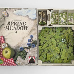 Op de afbeelding: De Spring Meadow bordspeldoos toont een rotsachtig landschap met een groene appel, een kompas en een rood geblokte doek. De open doos bevat een plastic sorteerbak gevuld met groene kartonnen puzzeltegels met een weide-thema.