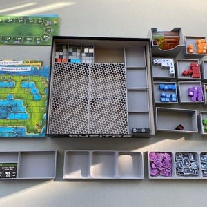 Könnte beinhalten: Ein grauer und schwarzer Spielorganizer mit mehreren Fächern zum Verstauen von Spielsteinen. Der Organizer hat ein Wabenmuster auf der Vorderseite. Die Spielsteine sind in verschiedenen Farben, darunter Rot, Blau, Grün und Gelb. Der Organizer ist für die Aufbewahrung des Spiels "The Great Run for Peat" konzipiert.