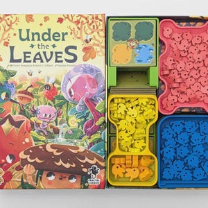Può includere: Un gioco da tavolo intitolato Under the Leaves con il coperchio della scatola che presenta illustrazioni colorate di una foresta e un interno aperto contenente vassoi in plastica verde, gialla e arancione pieni di piccoli gettoni in legno rossi, gialli, arancioni e blu.