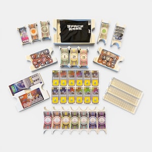 May include: A Space Base board game organizer set featuring white plastic trays for cards, dice, and tokens. Includes a black storage bag with the text SPACE BASE, multiple card slots, token containers, and individual player boards with colorful graphics.