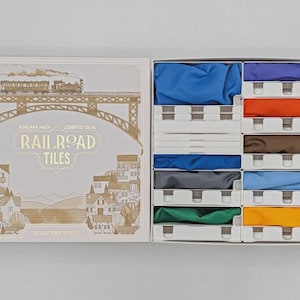 Puede incluir: Caja de juego de mesa "Railroad Tiles" abierta, con una ilustración de tren. Revela compartimentos con piezas de tela coloridas en azul, naranja, marrón, verde y amarillo. Es una edición de coleccionista.