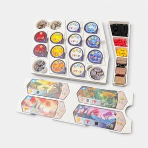 K&ouml;nnte beinhalten: Ein wei&szlig;er Kunststoff-Organizer f&uuml;r Brettspiele mit verschiedenen runden Spielsteinen in Rot, Gelb, Blau und Lila sowie einem separaten vertikalen Fach mit schwarzen, roten und gelben W&uuml;rfeln und Stapeln aus rechteckigen Papppl&auml;ttchen.